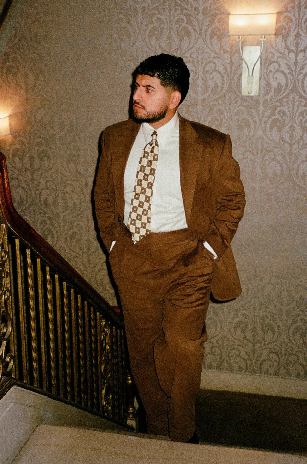 Julian in a brown suit and patterned tie on a staircase